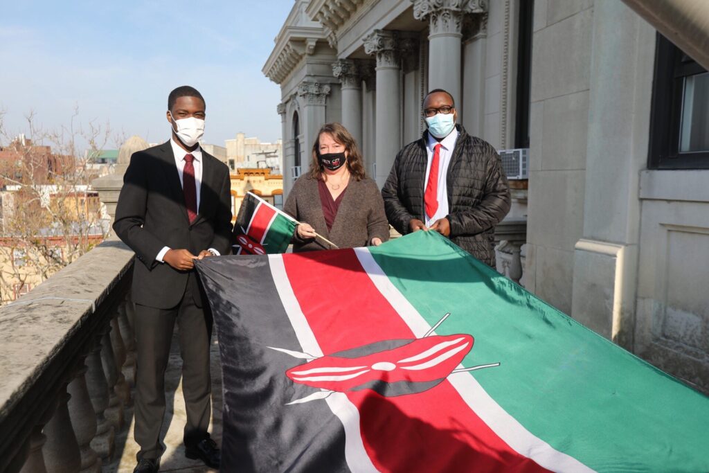 4 people holding the Kenayn Flag before it is raised. Alternation stripes of black, red, and green