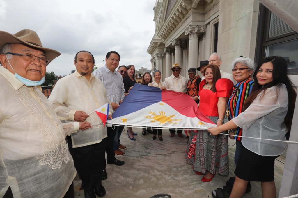 Mayor Fulop and a group of people hold the Philippine Flag before it is raised> It consists of a rectangular design that consists of a white equilateral triangle in which there is gold sun surrounded by gold stars. There is also a horizontal blue stripe followed by a horizontal red stripe