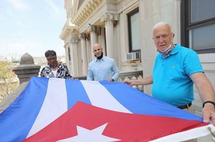  3 people holding the Cuban Flag before it is raised. The flag consists of five alternating stripes (three blue and two white) and a red equilateral triangle at the hoist, within which is a white five-pointed star.