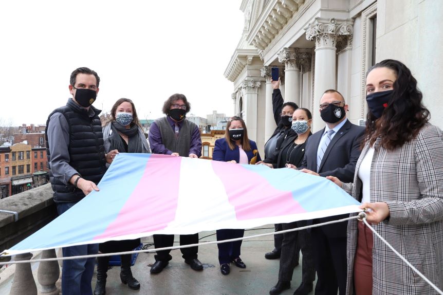 A group of people hold the flag before it is raised. The transgender flag contains the three colors represent baby blue (boys) baby pink (girls) and white for those who are transitioning, intersex or consider themselves as having an unidentified gender.