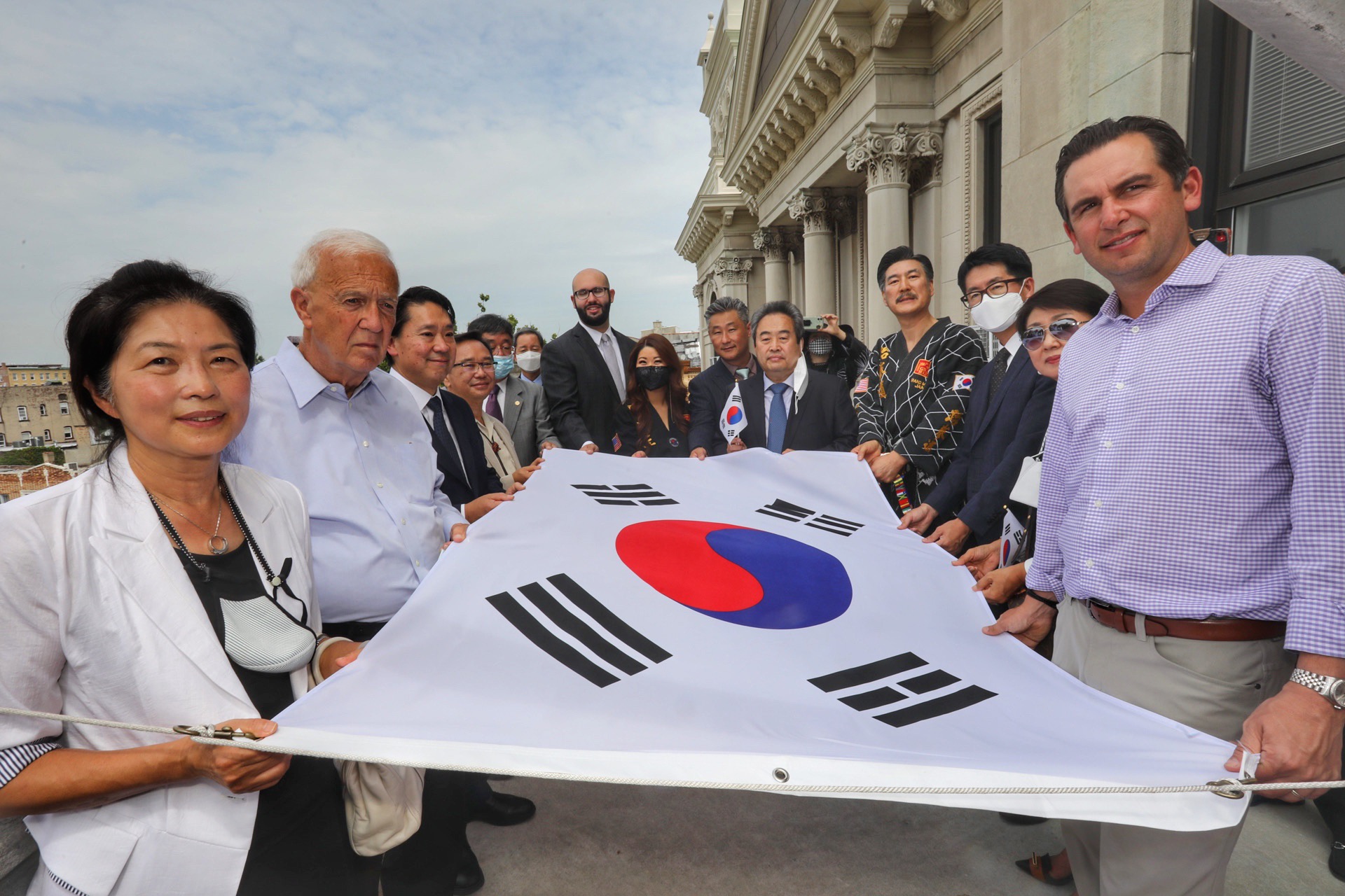 a group hold the Korean Flag before it is raisedThe Korean flag is called taegeukgi (pronounced teh-GUK-key). The colors of the flag are red, blue, and black on a white background. The flag stands for the three components of a nation: the land (the white background), the people (the red and blue circle), and the government (the four sets of black bars or trigrams)..