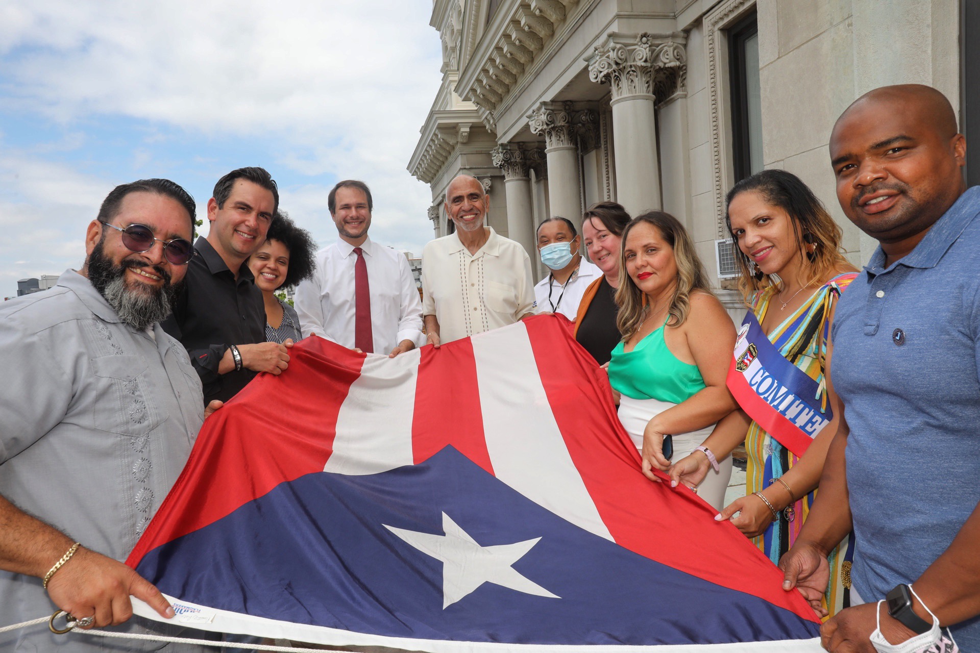A group of people hold the flag before it is raised. The three red stripes represent the blood from the brave warriors. The two white stripes represent the victory and peace that they would have after gaining independence. The white star represented the island of Puerto Rico. The blue represents the sky and blue coastal waters.
