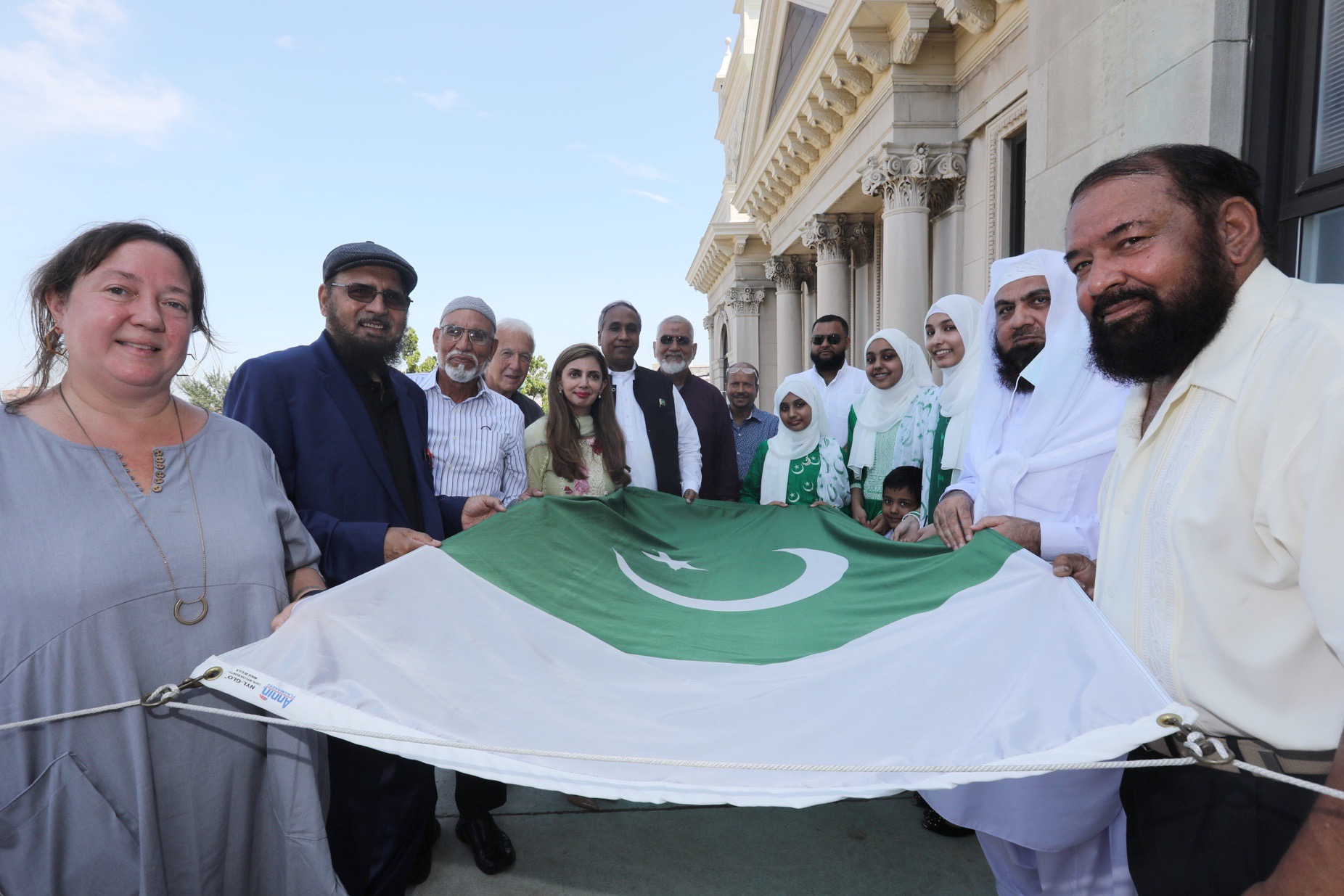 A group of people holding the Pakistan flag before it is raised. The City of Jersey City, Mayor Steven Fulop, Office of Municipal Council and Office of Cultural Affairs are proud to recognize the Muslim Council North America and the Muslim Federation of New Jersey as we celebrate the 74th Anniversary of Independence of Pakistan during the Flag Raising Ceremony on Friday, August 13, 2021. Pakistan, one of the two original successor states to British India, was partitioned along religious lines in 1947. Pakistan achieved its independence on August 14, 1947, with dominion status in the British Commonwealth of Nations. For almost 25 years following independence, it consisted of two separate regions, East and West Pakistan, now only made up of the western sector. The Pakistani community has directly contributed to the diversity and positive growth of Jersey City in various fields, including education, entrepreneurship, government, literature, drama, sculpture, music, food, and culture, as well as all aspects of life throughout the United States and abroad; and continues to influence our beliefs, embedded values and quest for independence. The City of Jersey City and the Pakistani community will proudly fly the flags of the United States and Pakistan high above City Hall in recognition of the cherished cultural and ethnic diversity of our community. It is also a reaffirmation of the long friendship built on common values and an abiding love for freedom..