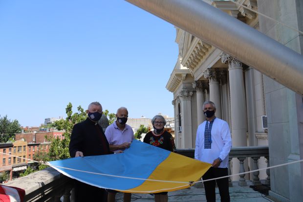 a group of peoplehold the flag before it is raised.The flag of Ukraine is a banner of two equally sized horizontal bands of blue and yellow. The combination of blue and yellow as a symbol of Ukrainian lands comes from the flag of the Kingdom of Galicia–Volhynia used in the 12th century. As a national flag, the blue and yellow bicolour has been officially used since the 1848 Spring of Nations, when it was hoisted over the Lviv Rathaus