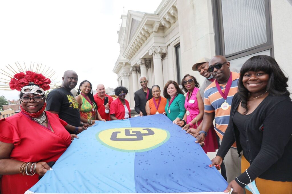 A group of people holding the flag before it is raised. The CARICOM colours are light blue representing the sky, and dark blue representing the sea of the Caribbean, yellow representing the sun, and green representing the vegetation of the region.