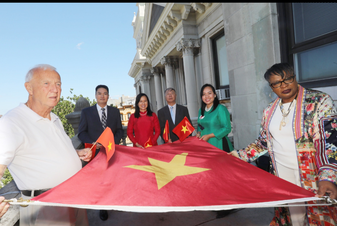 A group of people hold the flag prior to raising.The flag of Vietnam features a yellow five-pointed star on a red background. The flag is a symbol of the country's struggle against domination by the French and communist leadership. The star on the flag represents the country's national unity despite its turbulent past. This flag is the background for the flyer. Gold wordageappears detailing the event.