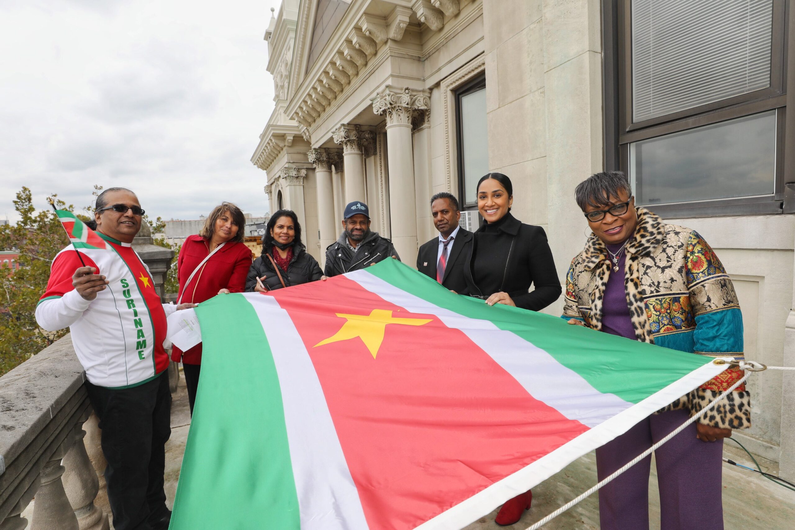 The Flyer has the Suriname Flag A group of people holding the flag prior to it being raised. The flag of Suriname is composed of five horizontal bands of green (top, double width), white, red (quadruple width), white, and green (double width) with a large, yellow, five-pointed star in the center.