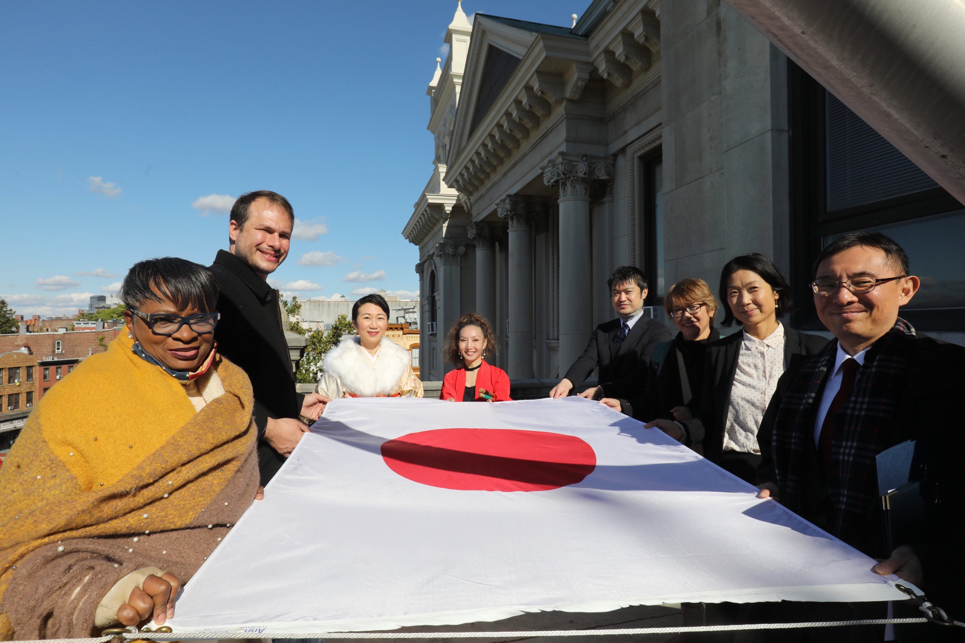 A group of people hold the flag before it is raised. The flag is a rectangular white banner bearing a crimson-red circle at its center.