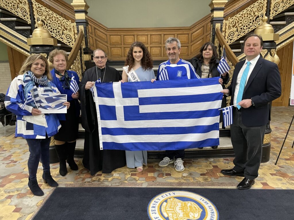 A group ofpeople hold the Greek Flag. The .flag is made of nine horizontal stripes that alternate between blue and white, and there is a white cross on a blue square in the top left.